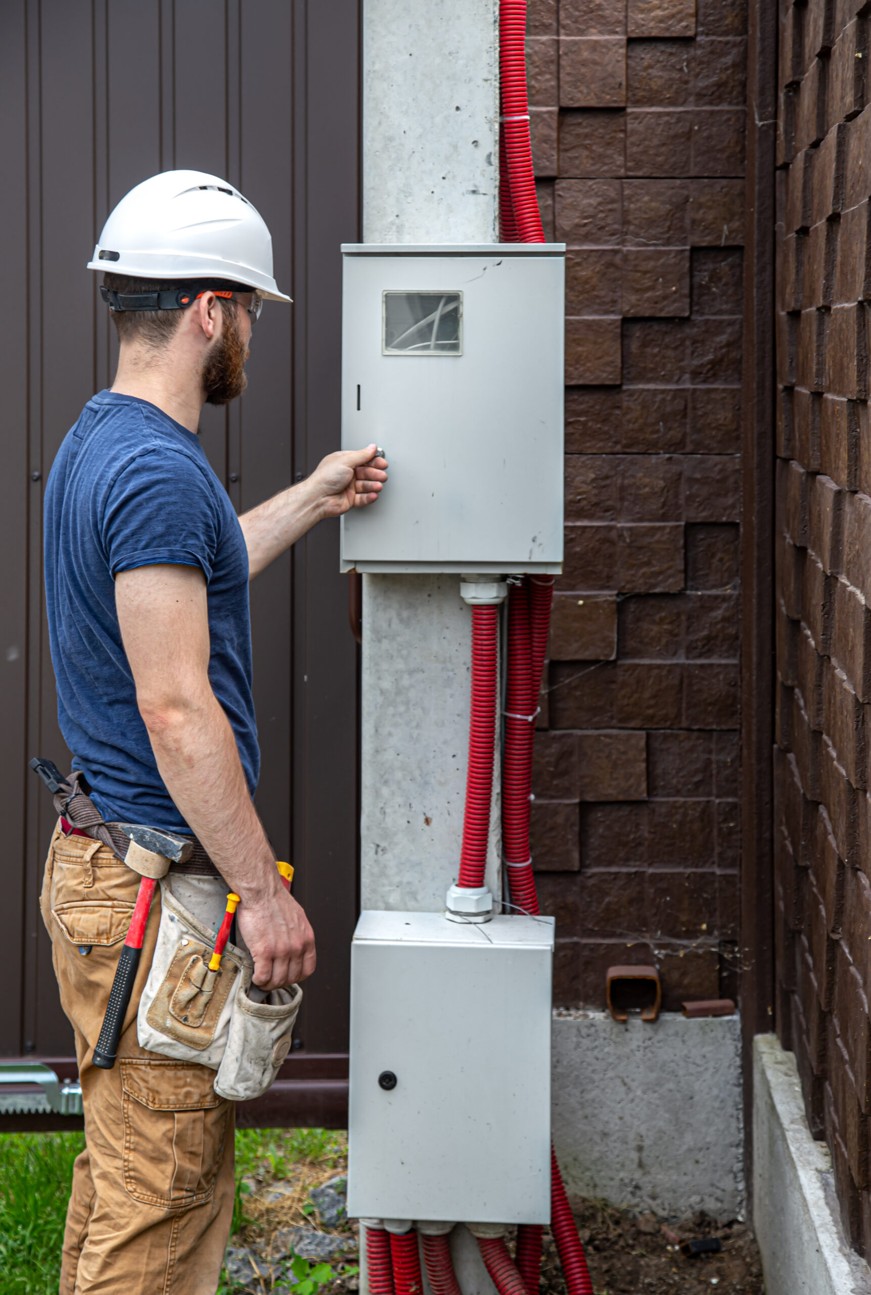 Electrician Builder at work, examines the cable connection in the electrical line in the fuselage of an industrial switchboard. Professional in overalls with an electrician’s tool.
