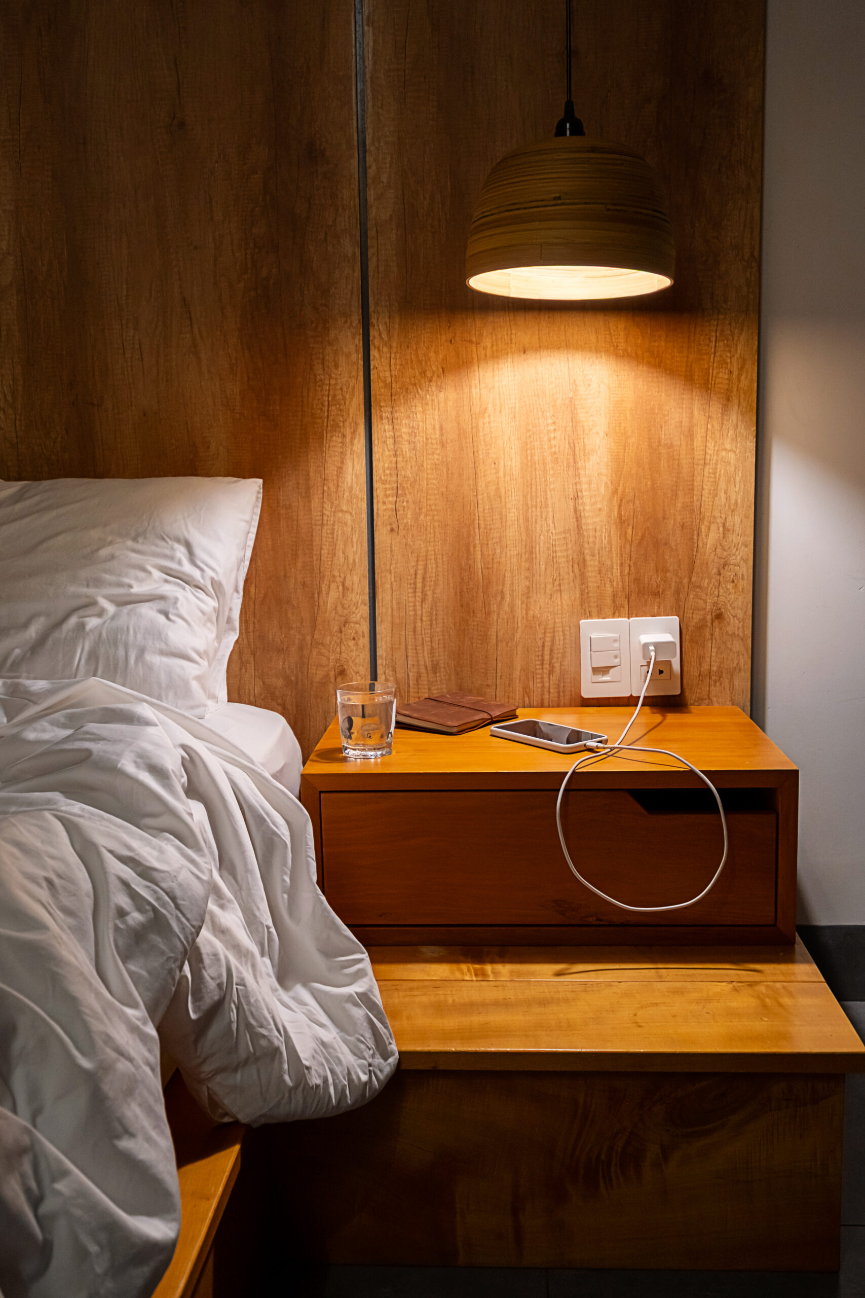 Empty bed with white pillow and sheet next to wooden nightstand and lamp.